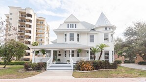 A white two-story house with a wrap-around porch, a turret, and a front yard with plants.