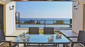 Mesa com tampo de vidro, cadeiras e tigelas, varanda com vista para o mar e corrimão branco.