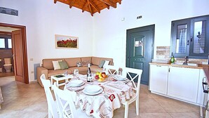 Uma área de refeições com mesa posta para quatro, um sofá, cozinha com armários e uma porta com sinalização.