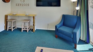A room with a blue armchair, a wooden desk with two stools, a flat-screen TV, and a 'WELCOME TO KEYLOIS' sign. - Casa Del Mar 118-welcome to Keylois (Galveston)