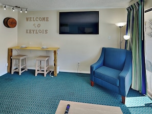 Ein Zimmer mit blauem Sessel, einem Holztisch mit zwei Hocker, einem Flachbildfernseher und einem Schild mit der Aufschrift 'WELCOME TO KEYLOIS'.