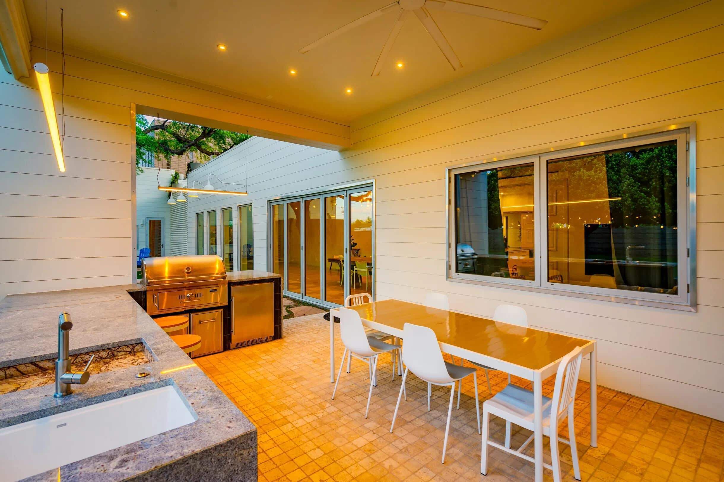 A modern kitchen with a built-in grill, a dining area with a glass table and chairs, and large windows offering a view of the outdoors.