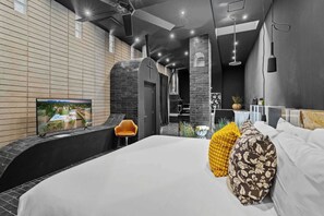 Ein modernes Hotelzimmer mit einem großen Bett, einem Fernseher links und einer kleinen Sitzecke mit einem Sessel.