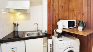 A compact kitchen with a sink, microwave, and washing machine, wooden walls, and a small shelf with a kettle and mug. - Cozy Studio in 13th Paris, Walk to Metro (Paris)