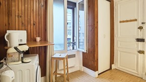 A small room with wooden paneling, a window with a view of buildings, a radiator, a white heater, a small wooden stool, and a white door with a handle. - Cozy Studio in 13th Paris, Walk to Metro (Paris)