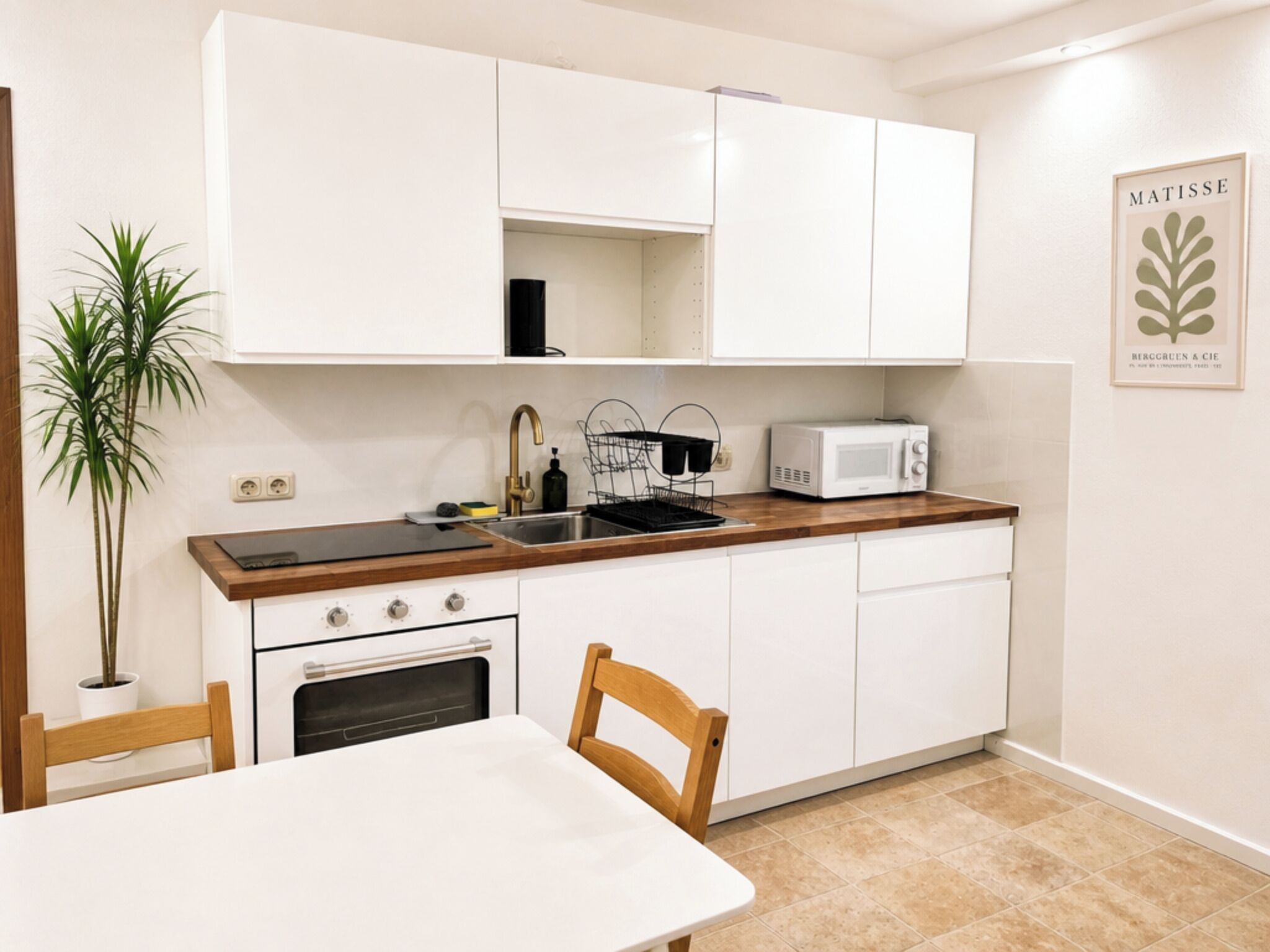 Une cuisine compacte avec des meubles blancs, un four à micro-ondes, un évier et une cuisinière. On y trouve une plante en pot et un tableau encadré accroché au mur.