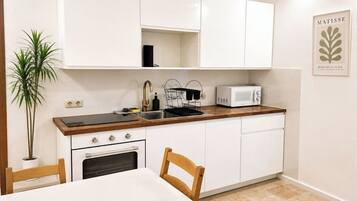 Une cuisine compacte avec des meubles blancs, un four à micro-ondes, un évier et une cuisinière. On y trouve une plante en pot et un tableau encadré accroché au mur.
