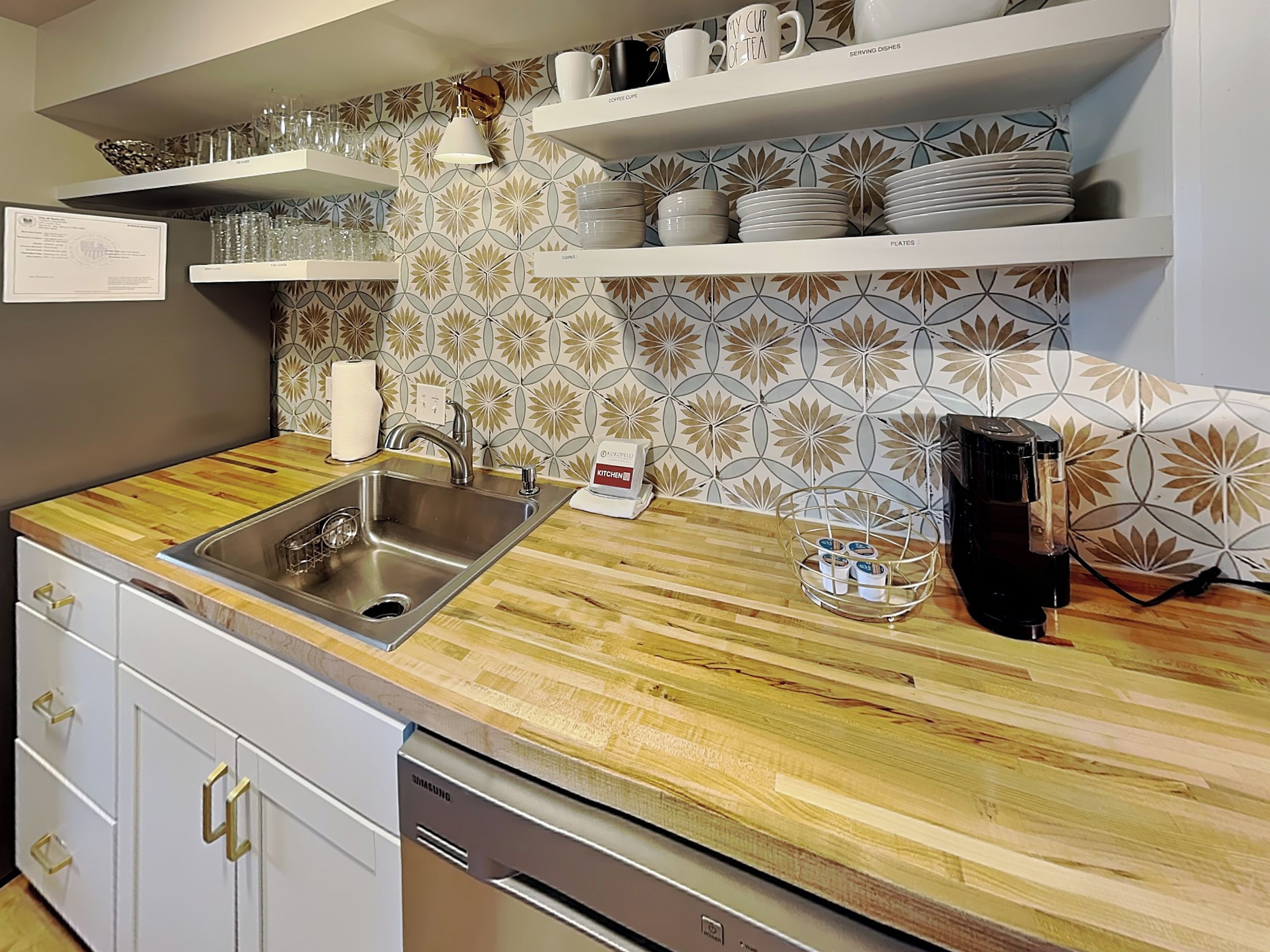 A modern kitchen with a stainless steel sink, wooden countertop, and built-in shelves holding serving dishes and coffee cups.