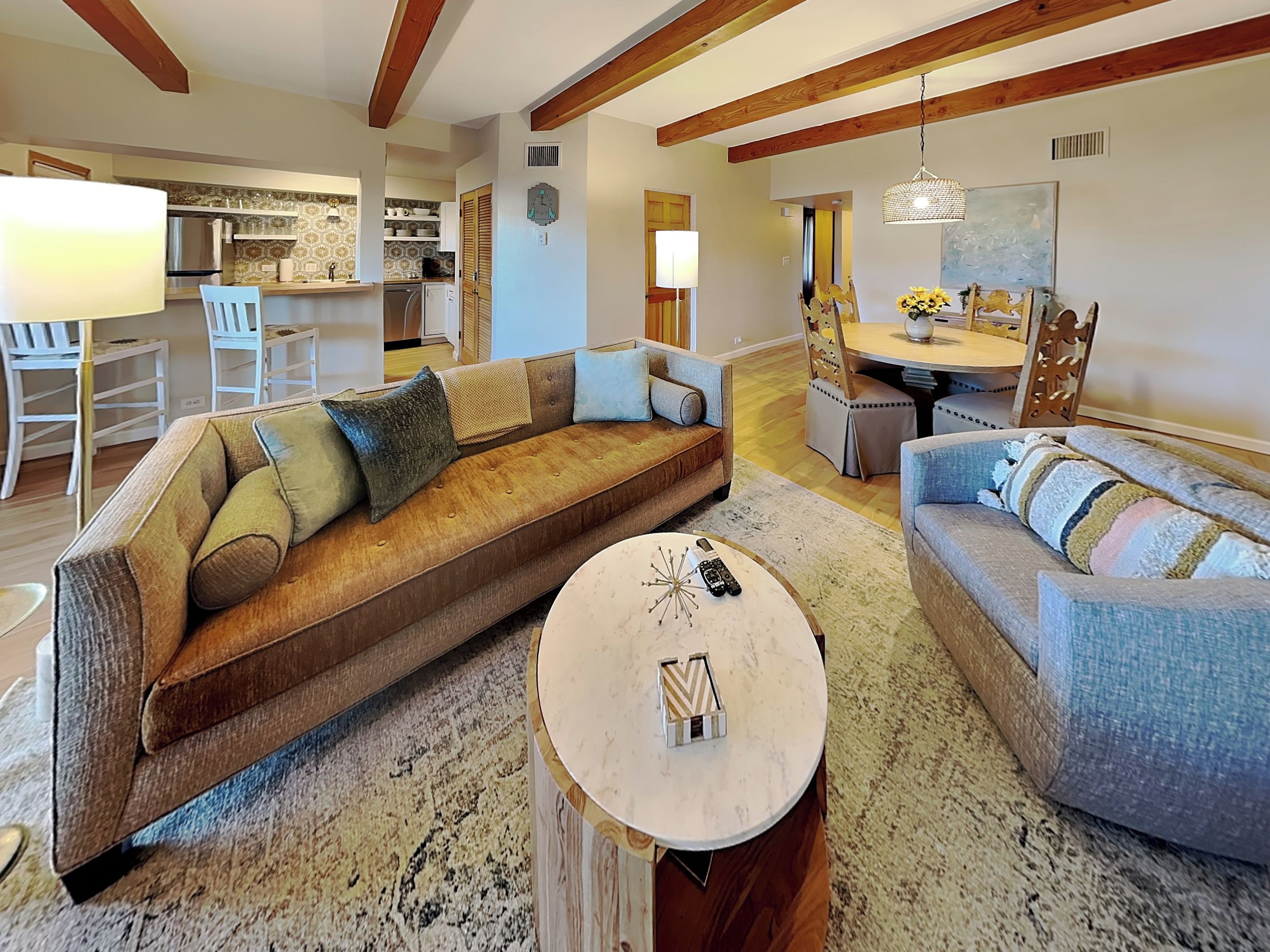 A spacious living room with a large brown sofa, a blue sofa, and a round coffee table. There is a kitchen in the background with a dining area.