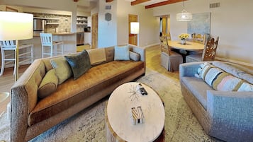 A spacious living room with a large brown sofa, a blue sofa, and a round coffee table. There is a kitchen in the background with a dining area.