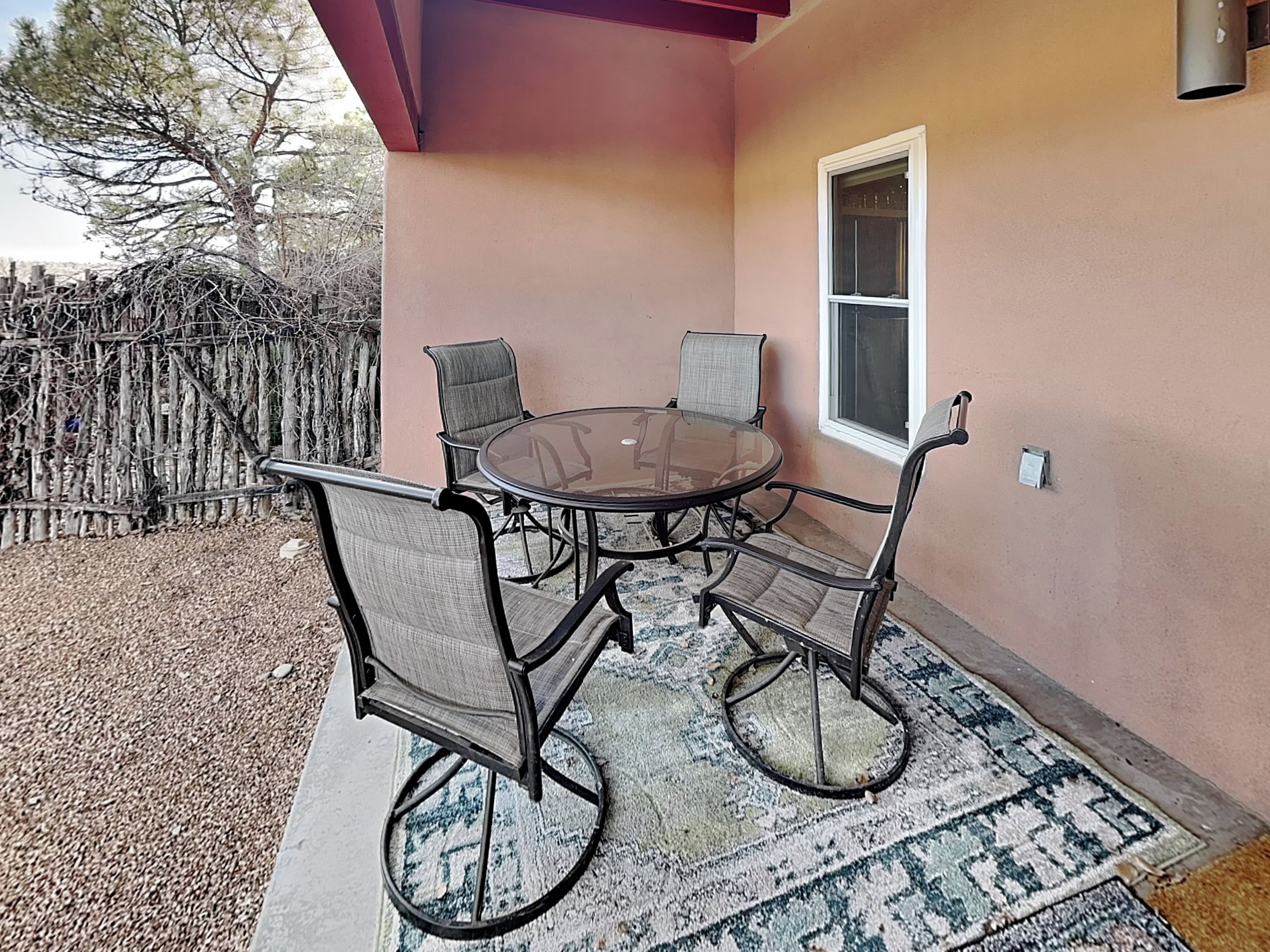 An outdoor patio with a round glass table and four chairs, a window, and a wooden fence in the background.