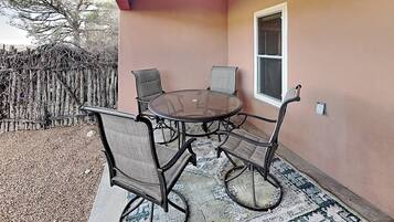 An outdoor patio with a round glass table and four chairs, a window, and a wooden fence in the background.