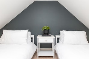 A bedroom with two white beds, a dark grey accent wall, a small white nightstand with a plant and books, and a carpeted floor. - 3BR Thames Retreat | Heathrow 10min | Train 5min (Egham)
