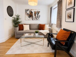 A living room with a grey sofa, a black leather chair, a glass coffee table, and a potted plant. - 3BR Thames Retreat | Heathrow 10min | Train 5min (Egham)