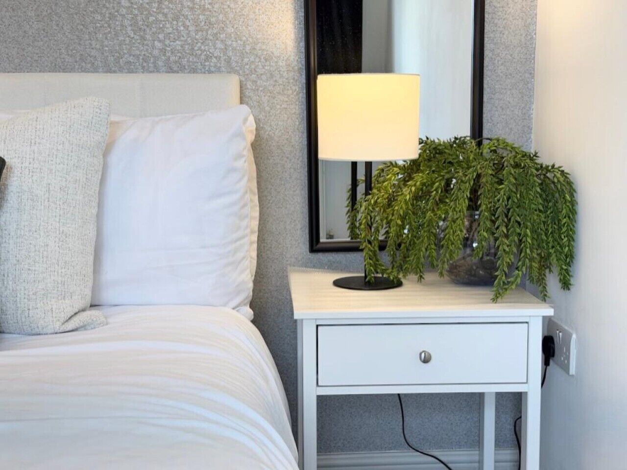 Una cama bien hecha con ropa de cama blanca, una mesita de noche blanca con un cajón, una lámpara y una planta en maceta.