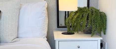 Una cama bien hecha con ropa de cama blanca, una mesita de noche blanca con un cajón, una lámpara y una planta en maceta.