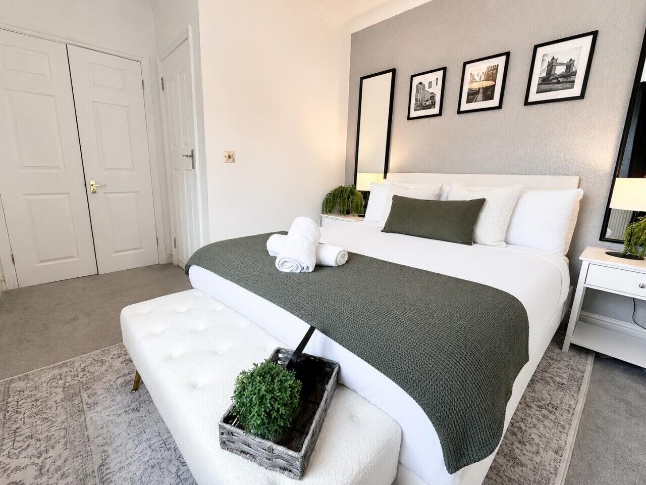 Un dormitorio con cama, una banca, una mesita de noche, una lámpara y un muro con cuadros.