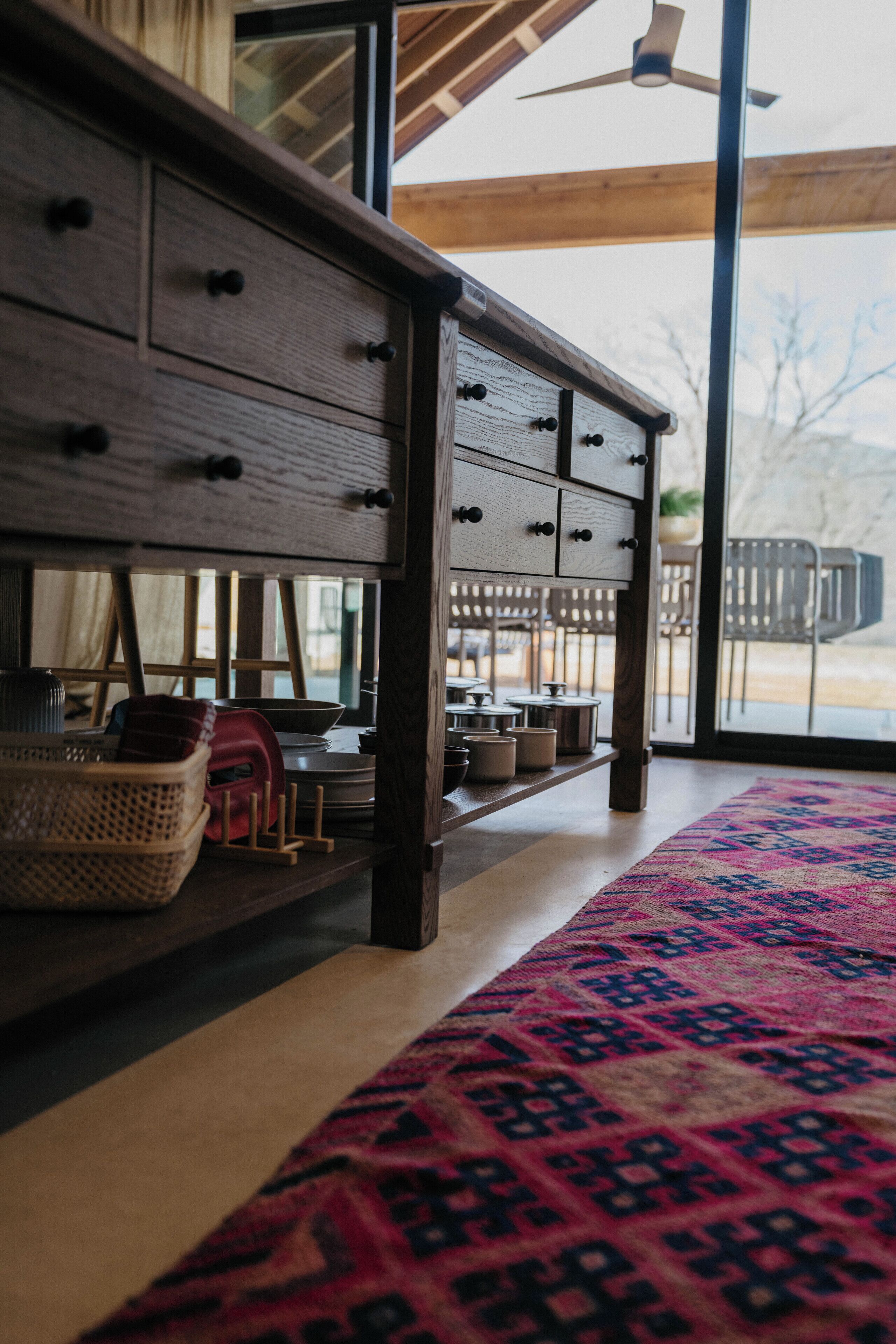 Een houten dressoir met lades, een kamerbreed tapijt met dessin en uitzicht op de omgeving door een groot raam.