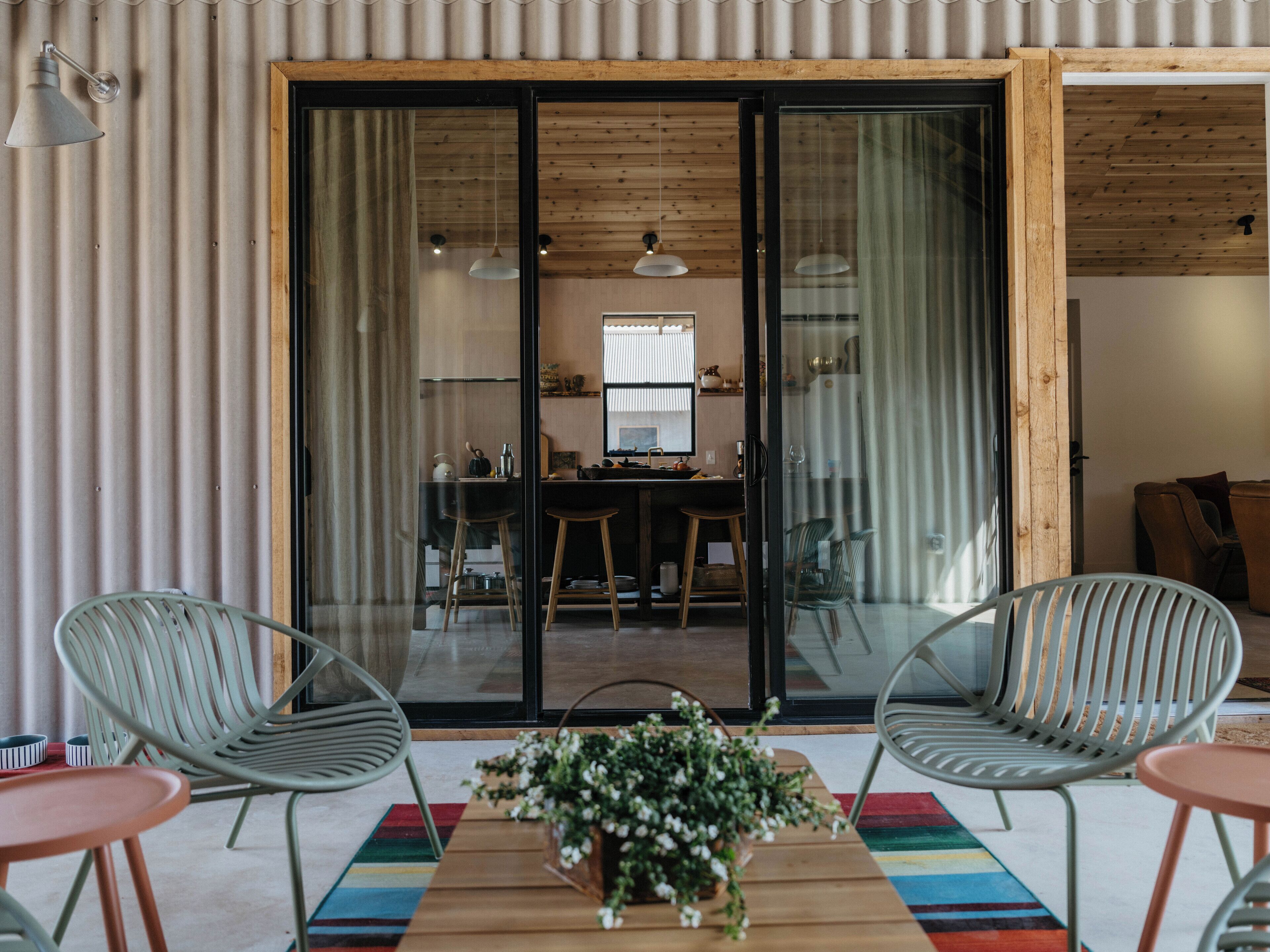 A modern interior with a wooden floor, a central table with a potted plant, two metal chairs, and a sliding glass door leading to a kitchen area.