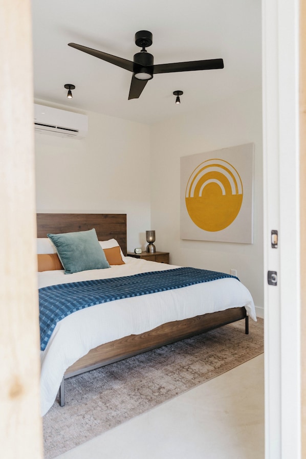 A bedroom with a bed, a ceiling fan, a wall-mounted air conditioner, and a piece of wall art. - Camp Cold Springs (Concan)