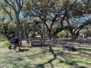 A picnic area with a grill, benches, and picnic tables surrounded by trees.