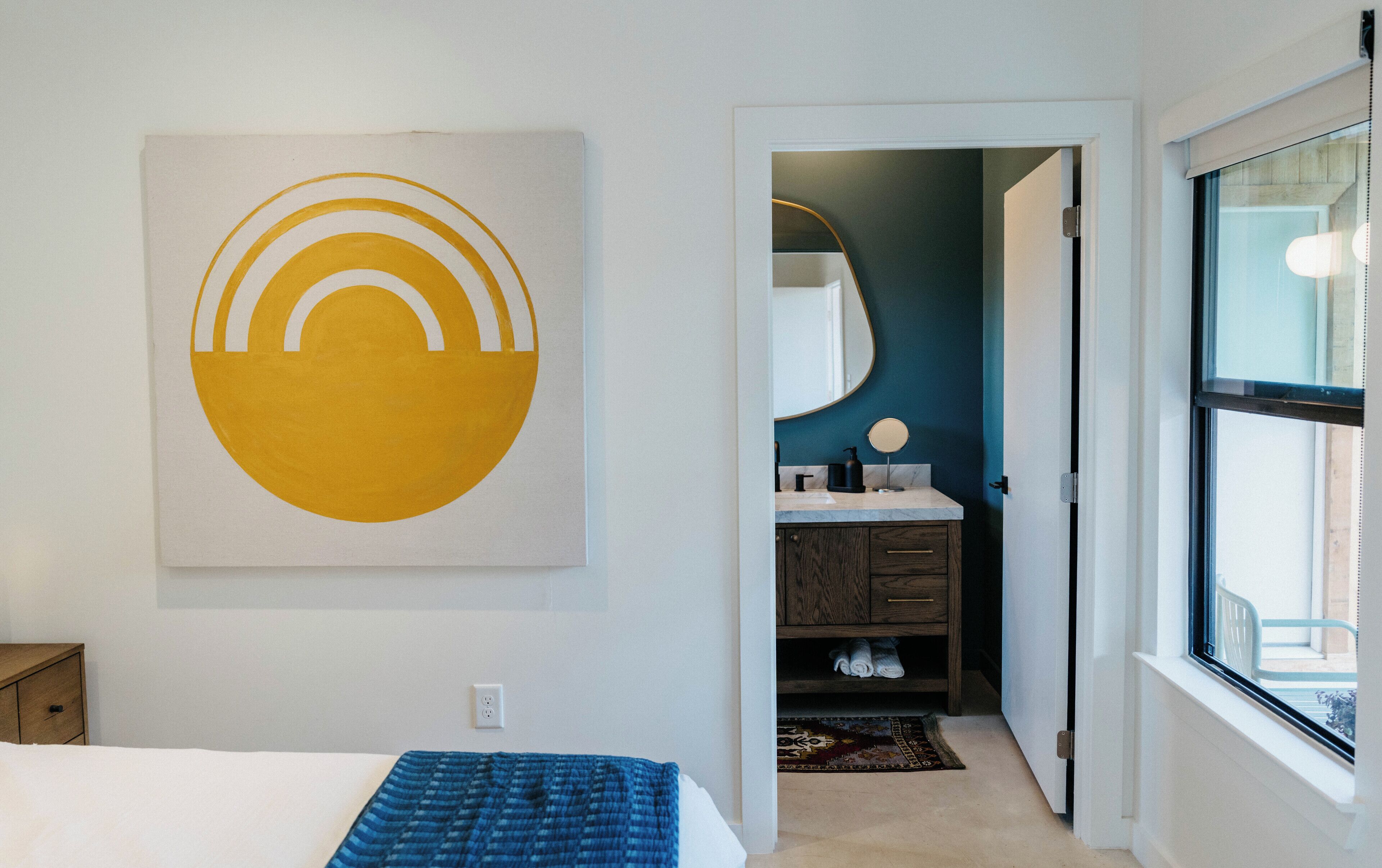 A bedroom with a bed, a wall-mounted artwork, a bathroom with a mirror and sink, and a view through a window.