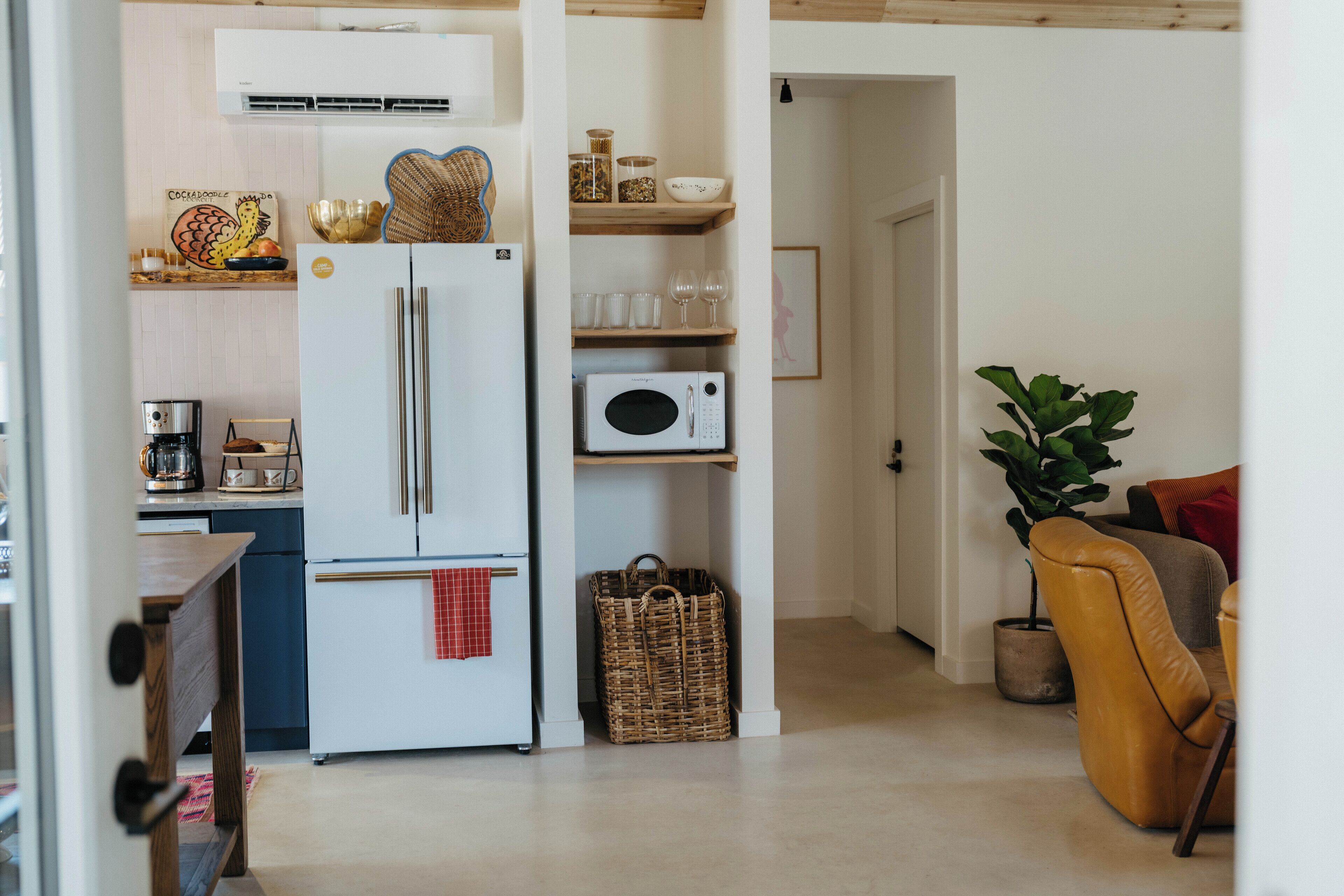 Eine moderne Küche mit einem weißen Kühlschrank, einer Mikrowelle und Holzregalen. Ein Korbwägelchen und ein Blumentopf stehen im Raum.