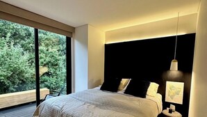 A modern bedroom with a large bed, a nightstand, a lamp, and a view of greenery outside.