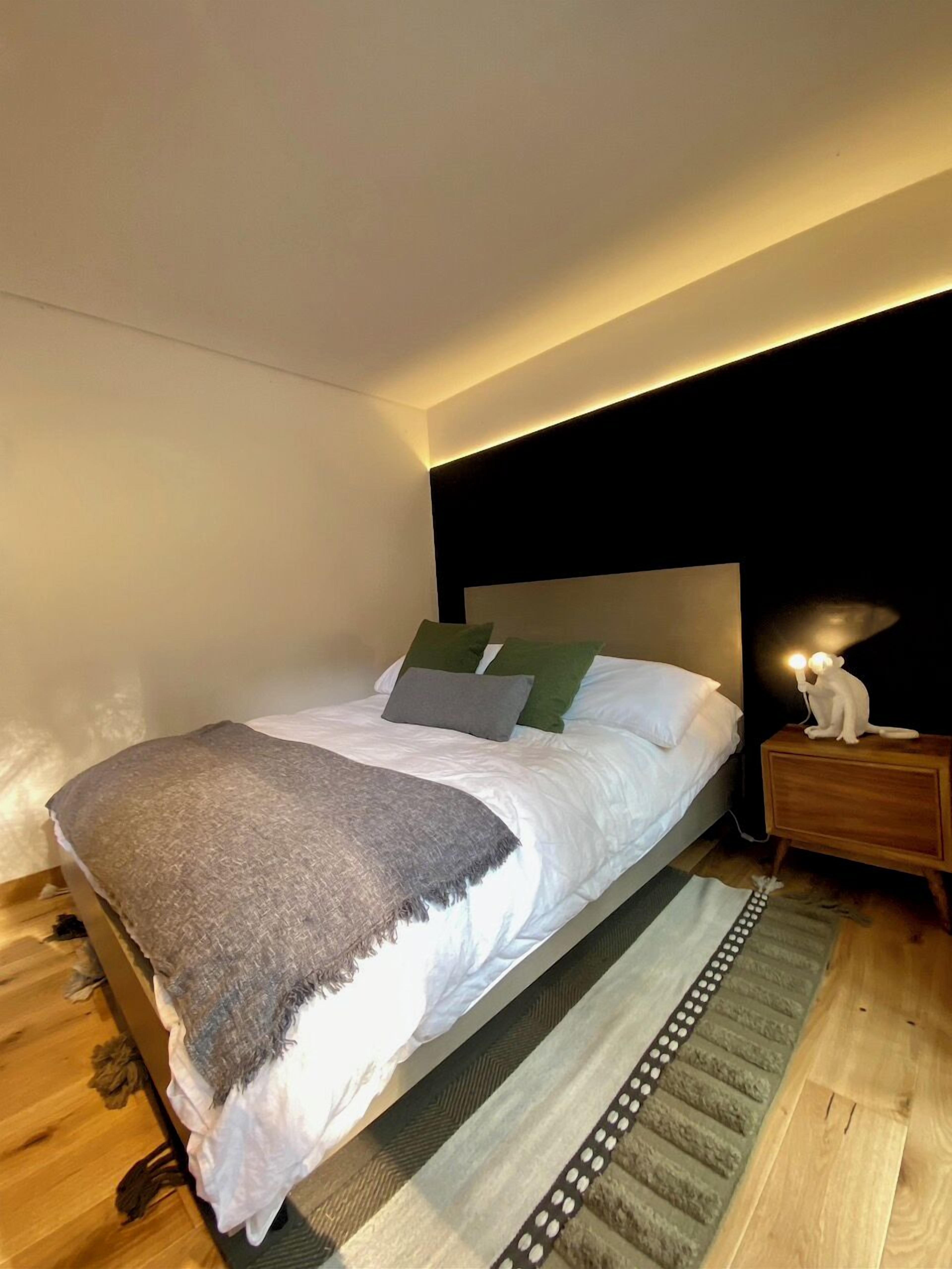 A modern bedroom with a large bed, a nightstand with a lamp, and a headboard with built-in lighting.