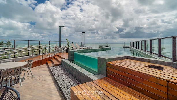 Una piscina infinita en la azotea con mobiliario de madera y barandilla de vidrio, con vistas al océano.