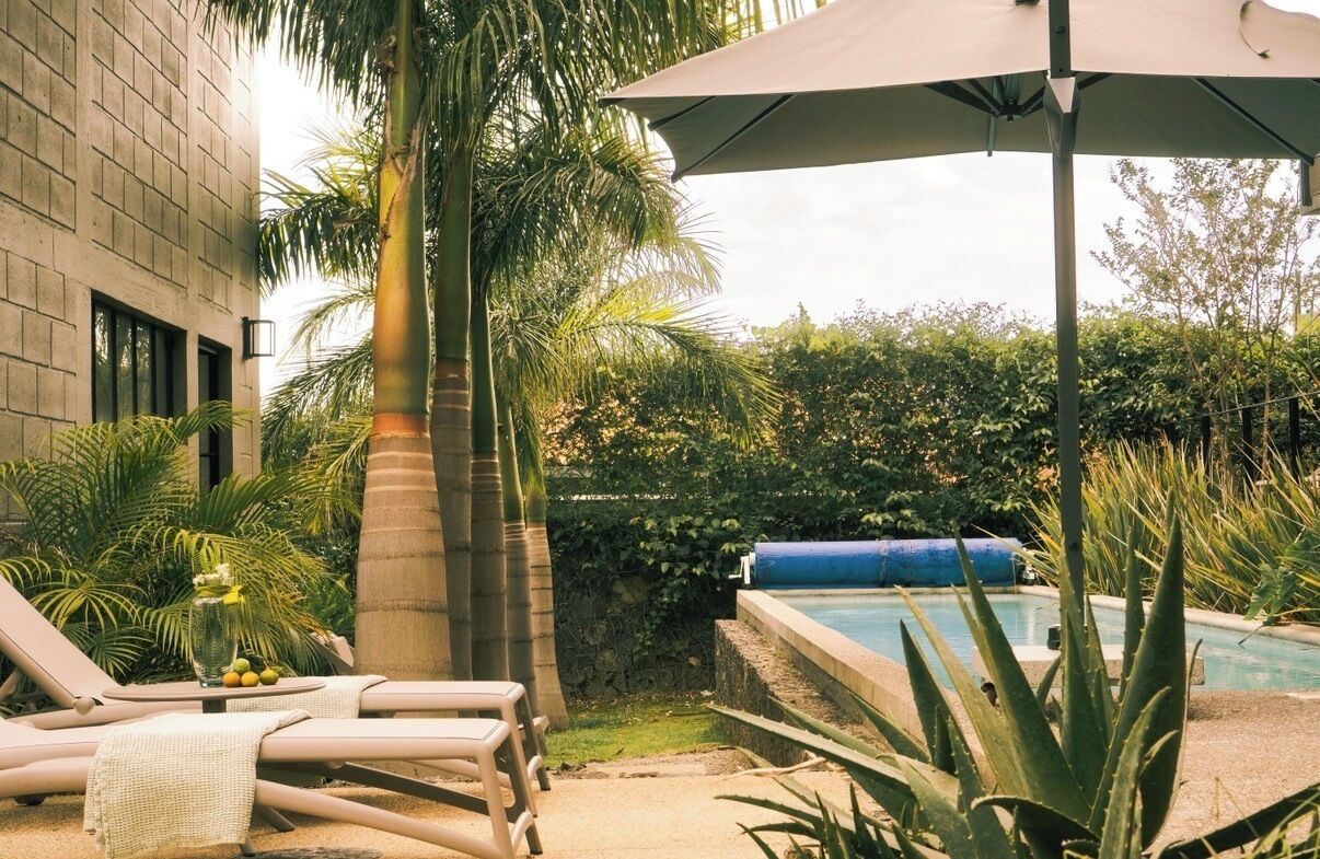 Un área de piscina con sillas de descanso y una mesa con frutas, rodeada de plantas tropicales y un muro de piedra.