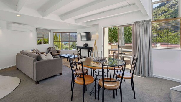 A modern living room with a sofa, dining table, and chairs, a TV, and large windows with a view of greenery. - Peaks & Cranbury 17B:  Location & Views ! (Queenstown)