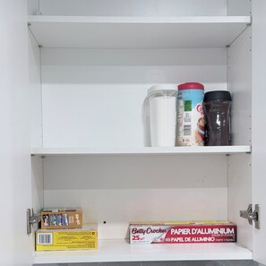Ein weißer Schrank mit zwei Einlegeböcken; der obere Boden enthält eine Kaffeeflühflasche und einen Thermosbehälter, während der untere Boden eine Betty Crocker Alufolie-Box beherbergt.