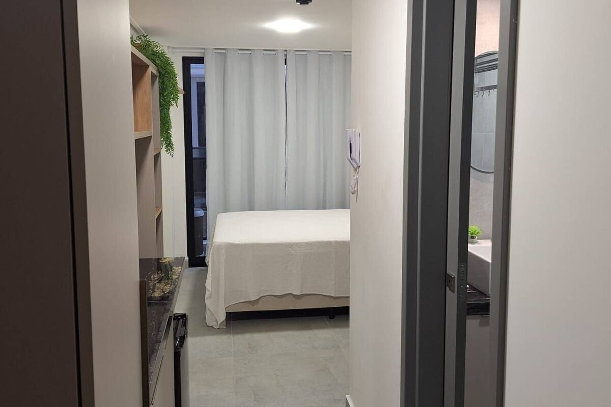 A narrow hallway leading to a bedroom with a bed, a small table, and a plant.