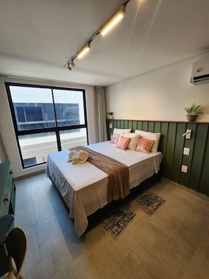 Habitación de hotel moderna con un ventanal grande, cama con cubrecama y almohadas color beige, cabecera verde, planta pequeña en maceta y aire acondicionado de pared.