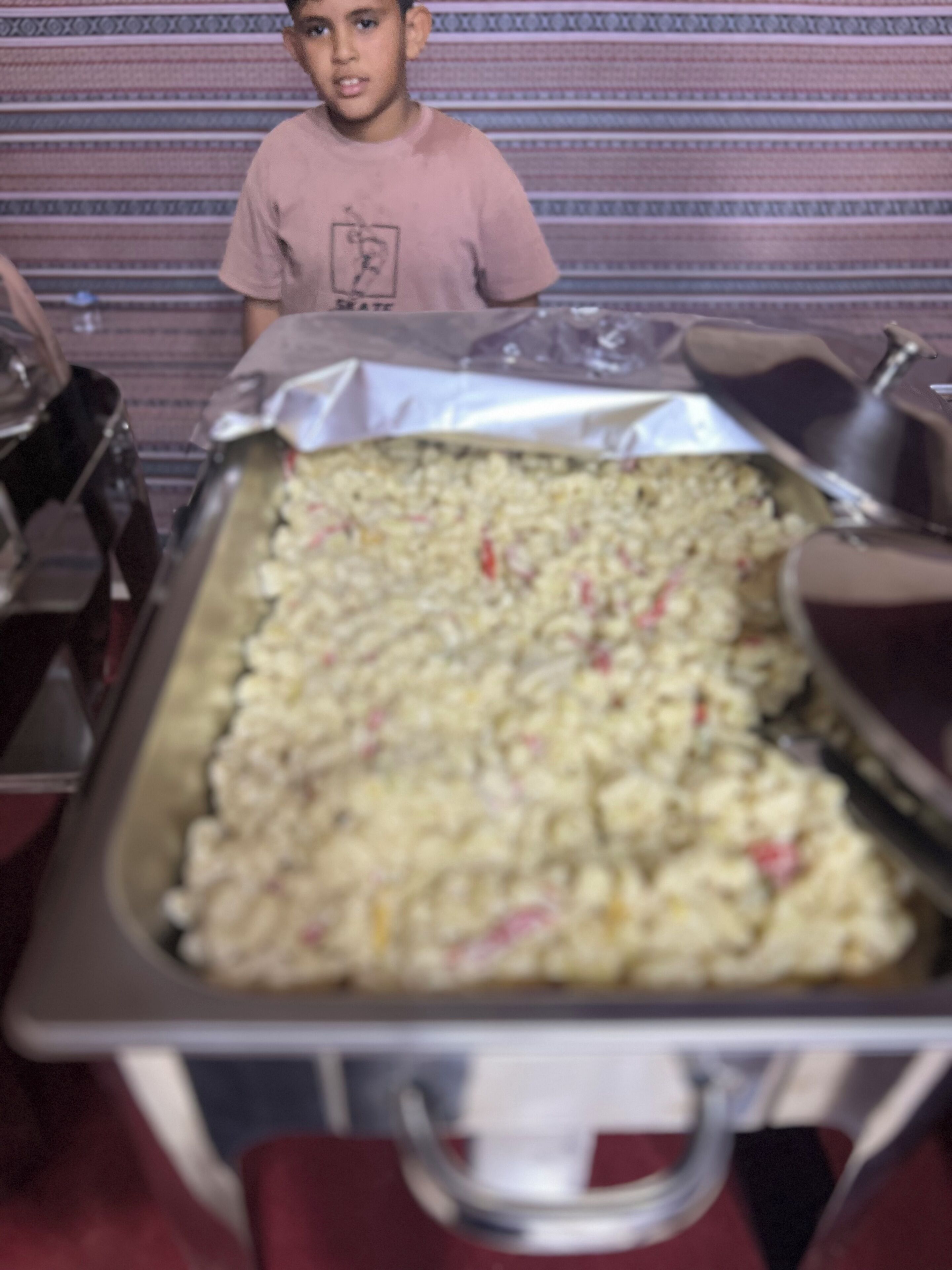 A buffet setup with a large tray of food, a person in a pink shirt, and a striped wall in the background.