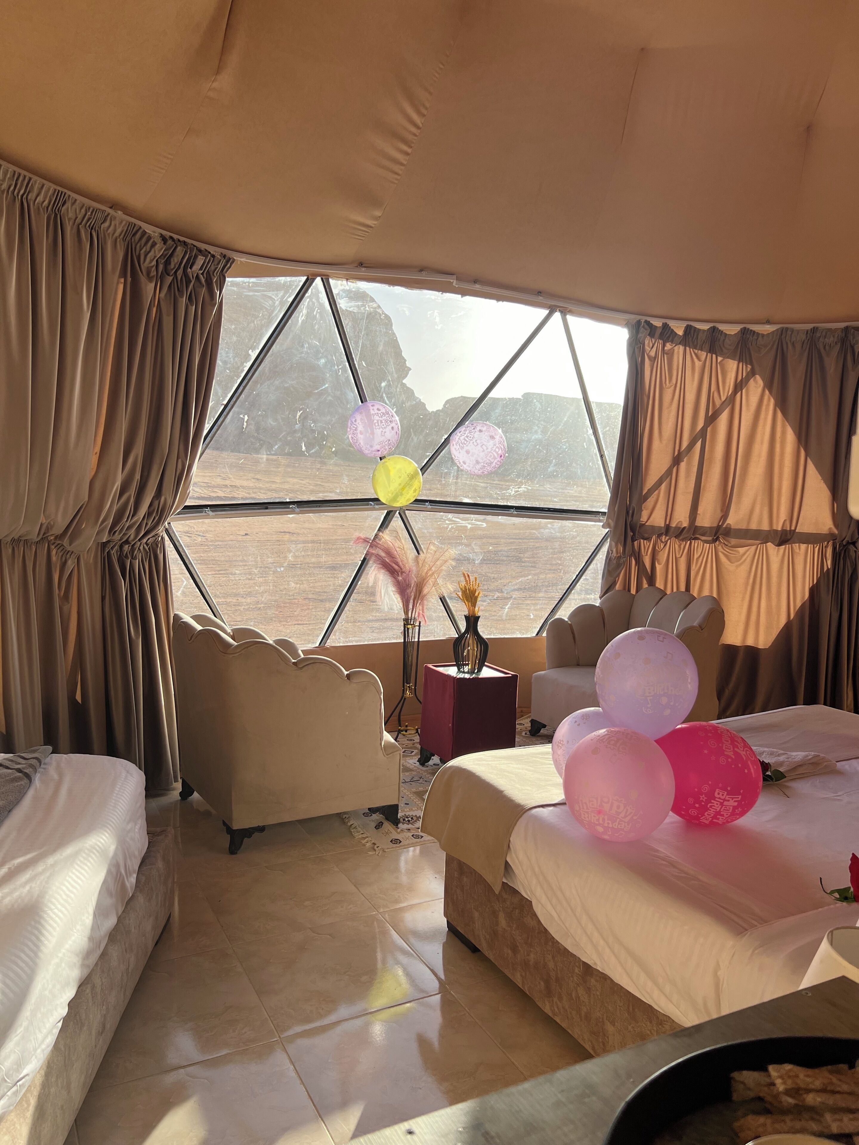 A room with a large window, two beds, a chair, and a small table with a vase.