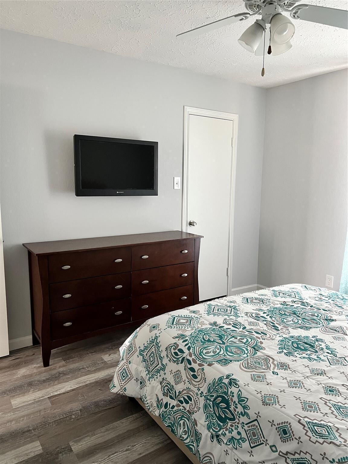 A bedroom with a wooden dresser, a flat-screen TV mounted on the wall, a door, and a patterned bedspread.