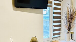 Quarto moderno e bem iluminado, com televisão de tela plana na parede, uma escrivaninha branca com um computador e uma xícara, uma cadeira e um radiador sob a escrivaninha.