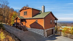 A two-story log cabin with a stone chimney, a covered porch, and a garage. - Valley Views | Hot Tub, Sauna, Game Room | Sleeps 14 (Sevierville)