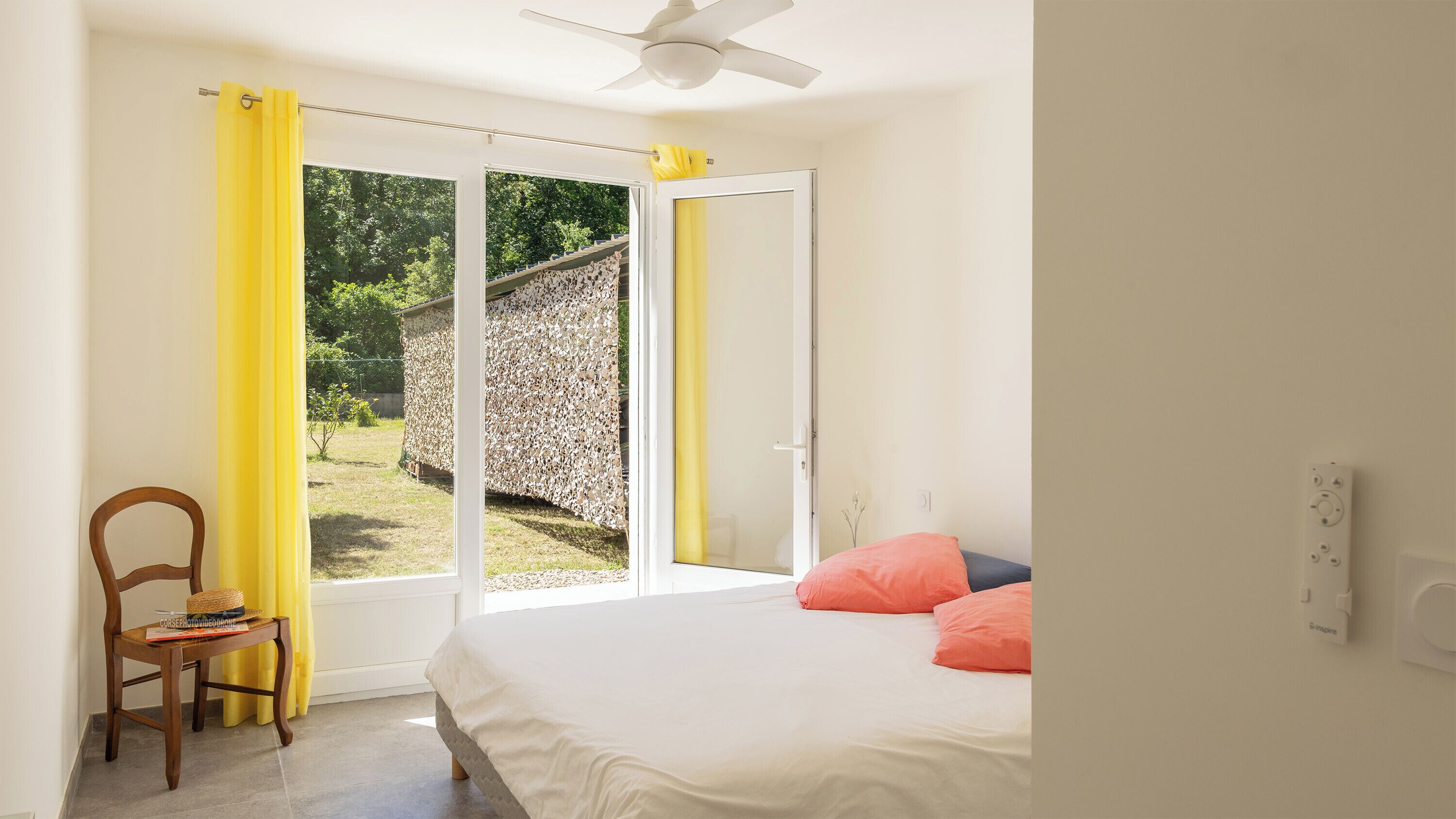 Une chambre à coucher avec un lit, une chaise, un ventilateur de plafond et une vue sur l’extérieur.