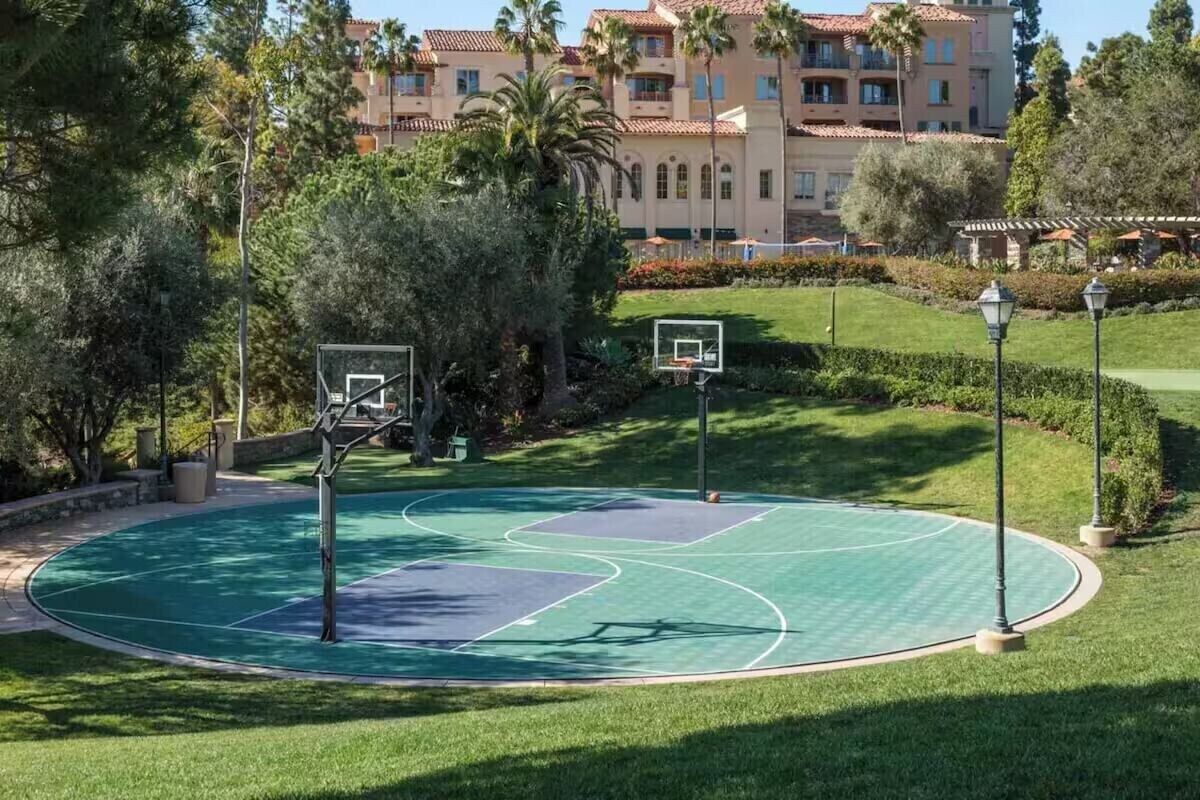 Een basketbalveld omringd door verzorgd gras en bomen, met een gebouw op de achtergrond.