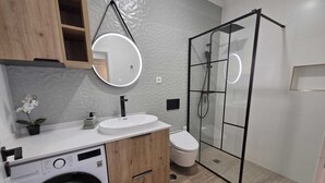 A modern bathroom with a built-in washing machine, a toilet, a glass-enclosed shower, and a round mirror with a light. - Cosy Apartment in Espiche near Praia da Luz (Luz)