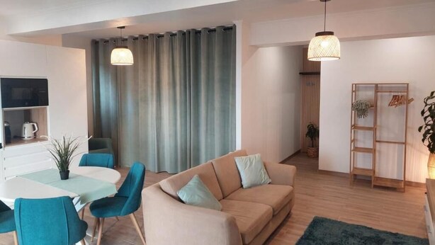 A modern living room with a beige sofa, teal chairs, a dining table, and a kitchen area with a TV and coffee maker. - Cosy Apartment in Espiche near Praia da Luz (Luz)