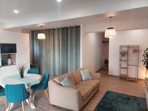 Una sala de estar moderna con un sofá color beige, sillas verde azulado, una mesa de comedor y una zona de cocina con televisión y cafetera.