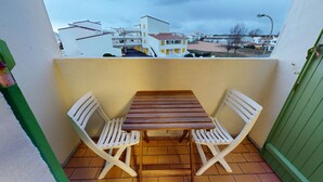 Een balkon met een houten tafel en twee witte stoelen, een betegelde vloer en een uitzicht op gebouwen in de verte.