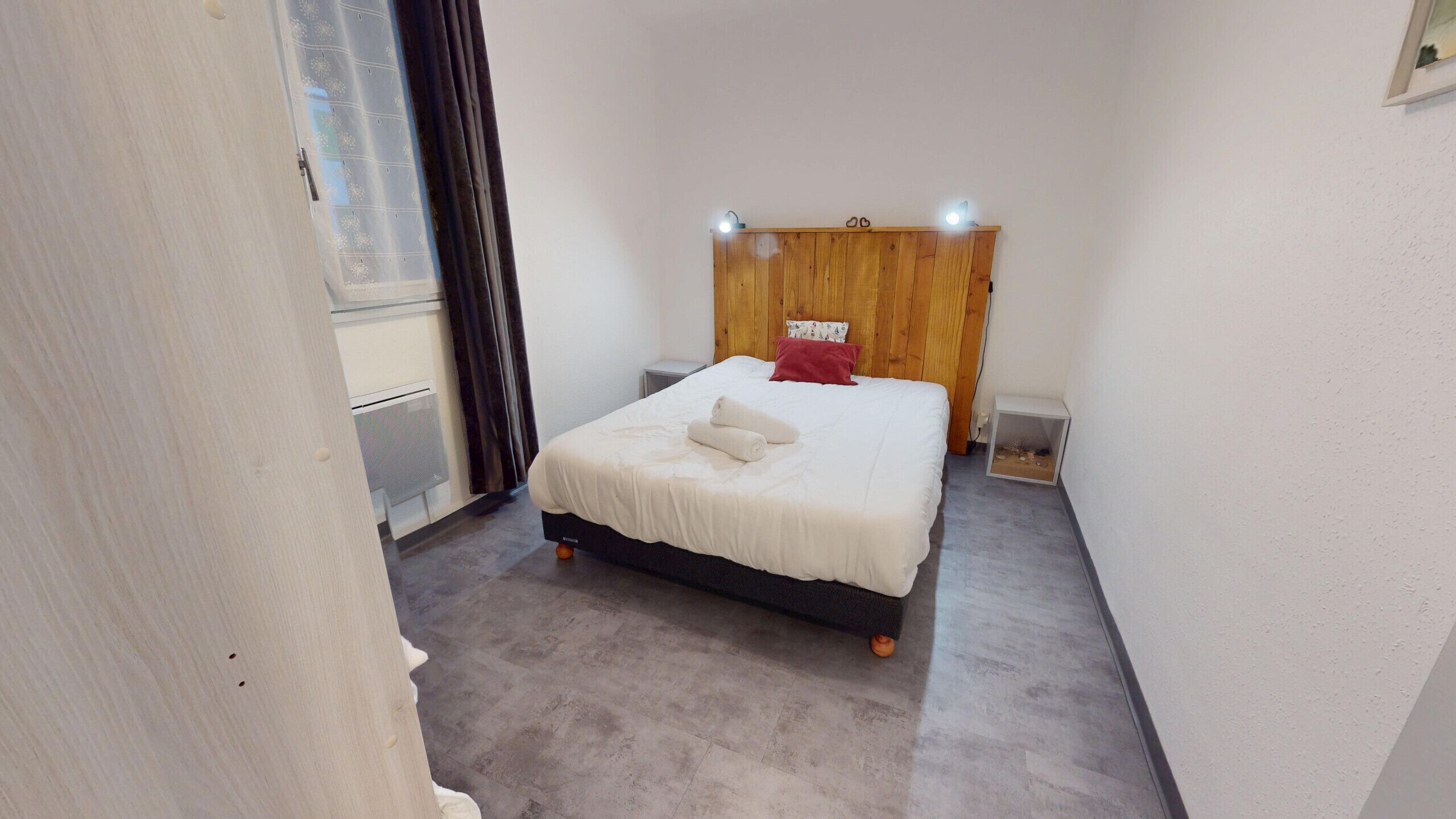Un dormitorio pequeño y ordenado con cabecera de madera, cama con ropa de cama blanca, almohada roja y una mesita de noche.