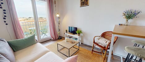 A cozy living room with a sofa, a coffee table, a TV on a white stand, a wicker chair, and a small dining area with a wooden table and bar stools.