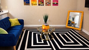 Una sala de estar con un sofá azul, una mesa de centro de vidrio con un jarrón, un tapete blanco y negro y una pared con carteles enmarcados.
