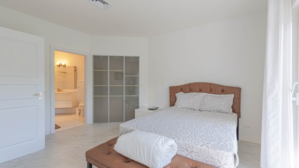 Ein Schlafzimmer mit Bett, einer Sitzbank, einem Kleiderschrank und einem durchgehenden Türrahmen einsehbaren Badezimmer.